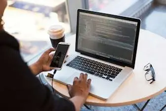 Mobile services thumbnail: a person in a coffee shop writing software on a laptop with a mobile phone connected to the computer.